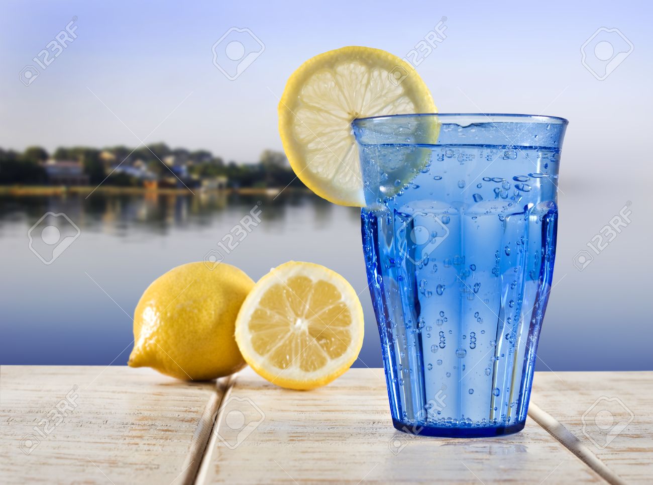 10762435-ein-blaues-Glas-mit-Mineralwasser-und-Zitrone-auf-einem-Holzdeck-mit-Blick-auf-das-ruhige-Wasser-ein-Lizenzfreie-Bilder